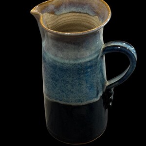May include: A handmade ceramic pitcher with a dark blue base, transitioning to lighter blue and tan hues towards the top. The pitcher has a curved handle and a spout for easy pouring. The pottery is glazed.