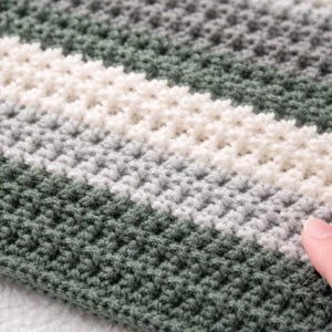 May include: Close-up of a hand-crocheted blanket with a textured stitch pattern. The blanket features horizontal stripes in shades of green, white, and gray. The soft, handmade textile is a cozy home decor item.