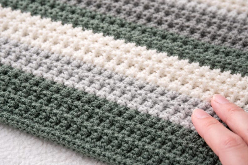 May include: Close-up of a hand-crocheted blanket with a textured stitch pattern. The blanket features horizontal stripes in shades of green, white, and gray. The soft, handmade textile is a cozy home decor item.