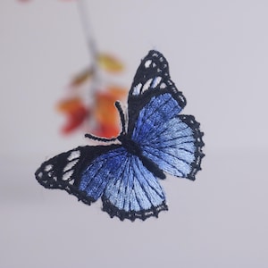 Puede incluir: Una mariposa bordada con alas azules y detalles en negro. Las alas están extendidas, mostrando las intrincadas puntadas. El fondo es un degradado suave y borroso de blanco y naranja, con toques de follaje.