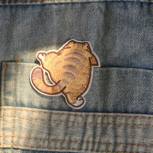 May include: Embroidered patch depicting a cartoon cat's rear end, in gold and brown hues, with a white border. The patch is positioned on a denim pocket, indicating it's for clothing or accessories.