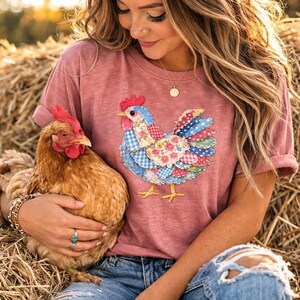 K&ouml;nnte beinhalten: Ein staubrosa T-Shirt mit einem farbenfrohen Patchwork-Hahn-Design. Der Hahn hat einen blauen K&ouml;rper, einen roten Kamm und mehrfarbige Fl&uuml;gel. Das Shirt wird mit einer Jeans getragen. Ein lebendes Huhn wird in den Armen gehalten.