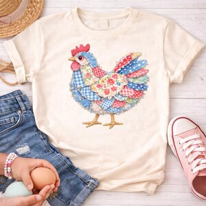 K&ouml;nnte beinhalten: Cremefarbenes T-Shirt mit einem bunten Patchwork-H&uuml;hnerdesign. Das Huhn hat einen roten Kamm und Kehllappen, mit blau, rot und floral gemusterten Federn. Das Shirt wird mit Jeansshorts und rosa Turnschuhen gezeigt.