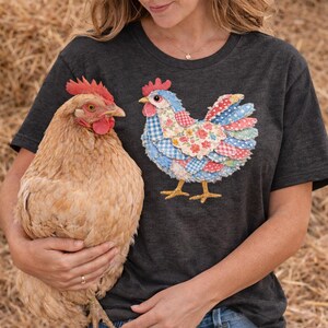 K&ouml;nnte beinhalten: Dunkelgraues T-Shirt mit einem farbenfrohen H&uuml;hnerdesign. Die Grafik zeigt ein Huhn im Patchwork-Stil mit blauen, roten und floralen Mustern. Ein echtes Huhn wird vor dem Shirt gehalten, was einen spielerischen Kontrast erzeugt.