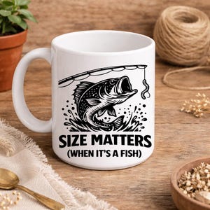 Może przedstawiać: Biały ceramiczny kubek z czarno-białą ilustracją ryby wyskakującej z wody, z wędką i robakiem. Tekst "SIZE MATTERS (WHEN IT'S A FISH)" jest wydrukowany poniżej.