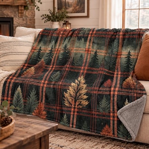 May include: A plush throw blanket featuring a dark green and orange plaid pattern overlaid with illustrations of evergreen trees. The blanket has a soft, light gray sherpa fleece lining visible at the folded corner and edges.