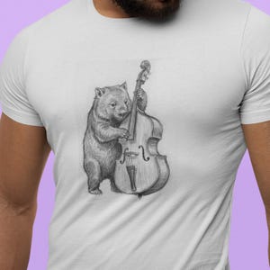 Puede incluir: Camiseta blanca con un dibujo a lápiz de un wombat tocando un contrabajo. El wombat está representado de forma realista, sosteniendo el instrumento. El diseño está centrado en la camiseta.