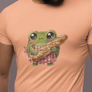 Puede incluir: Camiseta color melocotón con una ilustración caprichosa de una rana verde con un delantal a cuadros rojos y blancos, sosteniendo una baguette. La rana tiene ojos grandes y expresivos. El diseño está centrado en la camiseta.