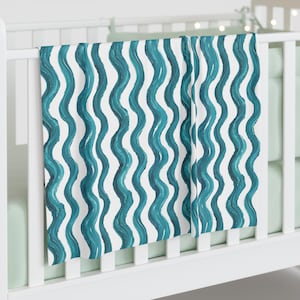 May include: A white baby crib with a teal and white patterned blanket draped over the side. The blanket features a repeating wavy design in shades of teal against a white background. The crib has a light green mattress.