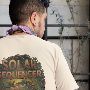 Può includere: T-shirt color crema con grafica circolare sul retro. La grafica presenta la scritta "SOLAR SEQUENCER" e un'illustrazione di un sintetizzatore, incorniciata da foglie verdi e piante grasse. La persona indossa una bandana viola.