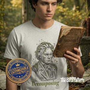 Pode incluir: Camiseta cinza claro com uma ilustração detalhada de Beethoven com aparência musgosa. O texto "Decomposing" está abaixo da imagem. Um emblema "Customizable" está à esquerda. A pessoa está segurando um livro antigo.