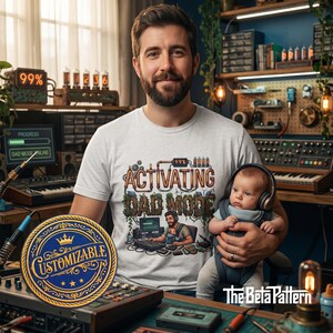 Könnte beinhalten: Hellgraues T-Shirt mit dem Text "ACTIVATING DAD MODE" in Braun- und Kupfertönen. Das Shirt zeigt eine Grafik eines Mannes am Computer. Ein Baby mit Kopfhörern wird von einem Mann gehalten. Ein anpassbarer Aufnäher befindet sich im Vordergrund.
