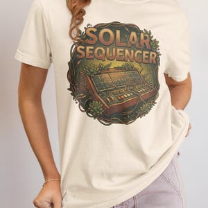 May include: Cream-colored t-shirt featuring a detailed graphic of a solar panel sequencer with the text "SOLAR SEQUENCER" in a circular design. The design is surrounded by botanical elements. The shirt has short sleeves.
