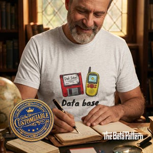 Op de afbeelding: Een lichtgrijs t-shirt met een cartoonontwerp met een rode floppy disk met het opschrift "Dad Jokes" en een gele mobiele telefoon. De woorden "Data base" staan onder de afbeeldingen. Een aanpasbare blauwe en gouden patch staat op de voorgrond.