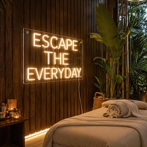 May include: A rectangular neon sign mounted on a dark wood paneled wall, displaying the illuminated text ESCAPE THE EVERYDAY in warm white light. The sign is positioned above a massage table covered with a light-colored cloth and a rolled towel.