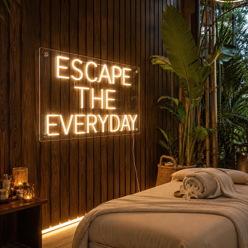 May include: A rectangular neon sign mounted on a dark wood paneled wall, displaying the illuminated text ESCAPE THE EVERYDAY in warm white light. The sign is positioned above a massage table covered with a light-colored cloth and a rolled towel.
