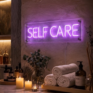 May include: A rectangular neon sign mounted on a textured wall, displaying the words SELF CARE in bright purple light. The sign is secured with four silver metal standoffs at the corners and features a clear acrylic backing with a visible power cord.