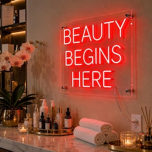 May include: A rectangular clear acrylic sign with bright red neon lettering that reads BEAUTY BEGINS HERE. The sign is mounted with four silver hardware standoffs and is illuminated against a wall, featuring a power cord extending to a wall outlet.