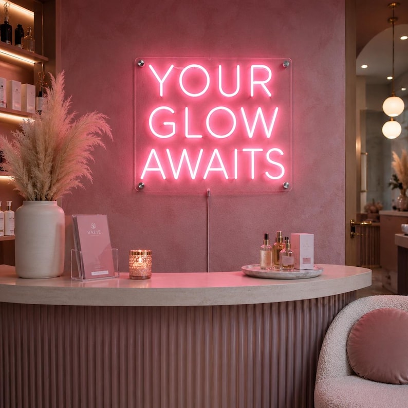 May include: A rectangular neon sign mounted on a pink wall with the text YOUR GLOW AWAITS in bright pink light. The sign is secured with four silver hardware standoffs at the corners and features a clear acrylic backing with a visible power cord.