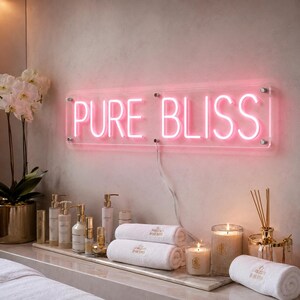 May include: A rectangular clear acrylic sign with pink neon lettering that reads PURE BLISS. The sign is mounted on a wall above a collection of white towels, glass bottles, and lit candles in a spa-like setting with a white orchid plant.