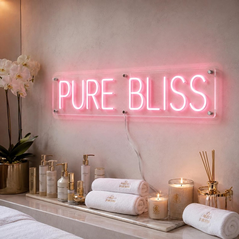 May include: A rectangular clear acrylic sign with pink neon lettering that reads PURE BLISS. The sign is mounted on a wall above a collection of white towels, glass bottles, and lit candles in a spa-like setting with a white orchid plant.