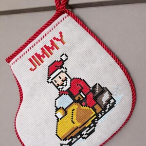 May include: A handmade Christmas stocking with a pixelated Santa Claus riding a yellow snowmobile. The stocking is white with a red trim and a red hanging loop. The name "JIMMY" is embroidered in red letters.