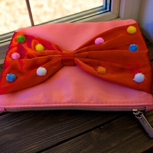 Könnte beinhalten: Eine rosa Clutch mit einer großen orangefarbenen Schleife, die mit bunten Pompons verziert ist. Die Clutch hat einen Reißverschluss und ein kleines Etikett am Reißverschlussanhänger. Die Schleife ist der Blickfang und verleiht dem Accessoire ein verspieltes und dekoratives Element.