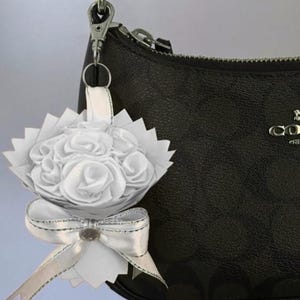 May include: A white floral bouquet charm with a silver bow and a silver clasp, attached to a black handbag. The bouquet features white fabric roses and a decorative bow with a silver trim.