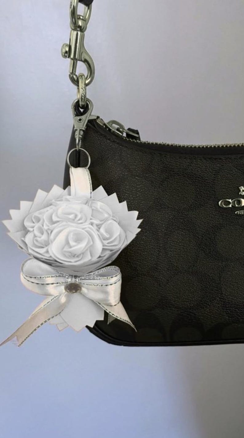 May include: A white floral bouquet charm with a silver bow and a silver clasp, attached to a black handbag. The bouquet features white fabric roses and a decorative bow with a silver trim.