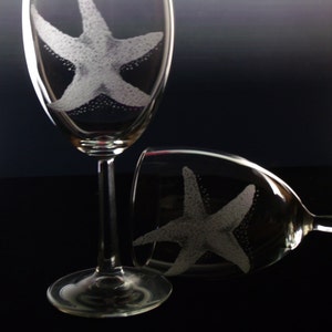 Peut inclure: Deux verres &agrave; vin en verre transparent avec des motifs d'&eacute;toiles de mer grav&eacute;s. Les verres sont sur une surface noire.