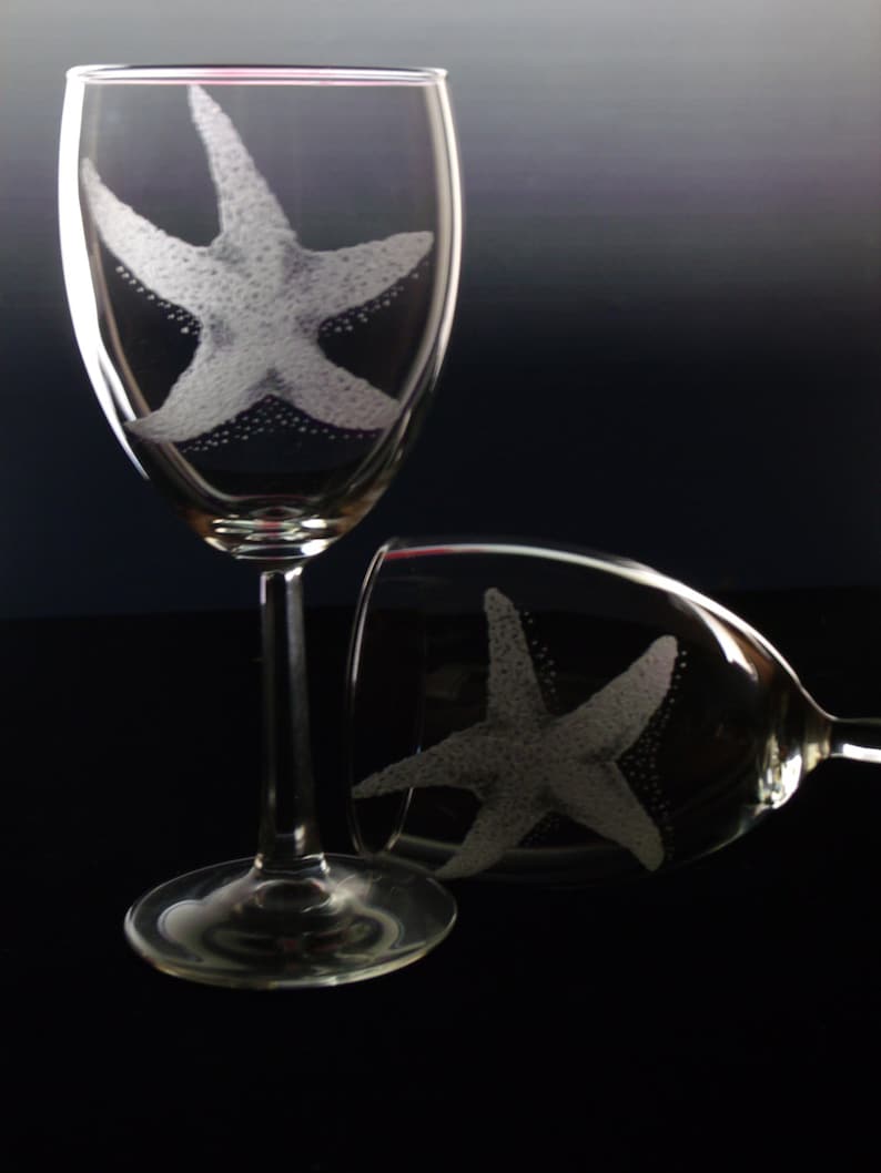 Peut inclure: Deux verres &agrave; vin en verre transparent avec des motifs d'&eacute;toiles de mer grav&eacute;s. Les verres sont sur une surface noire.