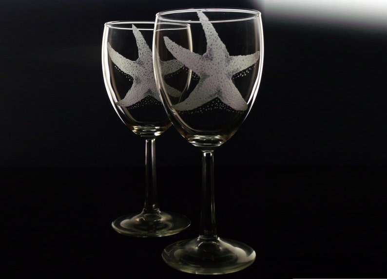Peut inclure: Deux verres &agrave; vin transparents avec des motifs d'&eacute;toiles de mer grav&eacute;s. Les verres sont sur un fond noir.