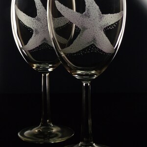 Peut inclure: Deux verres &agrave; vin en verre transparent avec des &eacute;toiles de mer blanches grav&eacute;es. Les verres sont sur un fond noir.