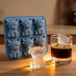 May include: A blue silicone ice cube tray with six Highland cow-shaped molds. A clear ice cube cow sits on a wooden surface next to a glass of dark liquid with a cow-shaped ice cube. The ice cubes have detailed features.