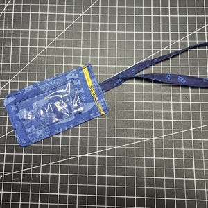 May include: Blue fabric ID badge holder with a clear plastic window and a matching lanyard. The holder has a yellow zipper and a blue patterned design. The lanyard is also blue with a repeating pattern.