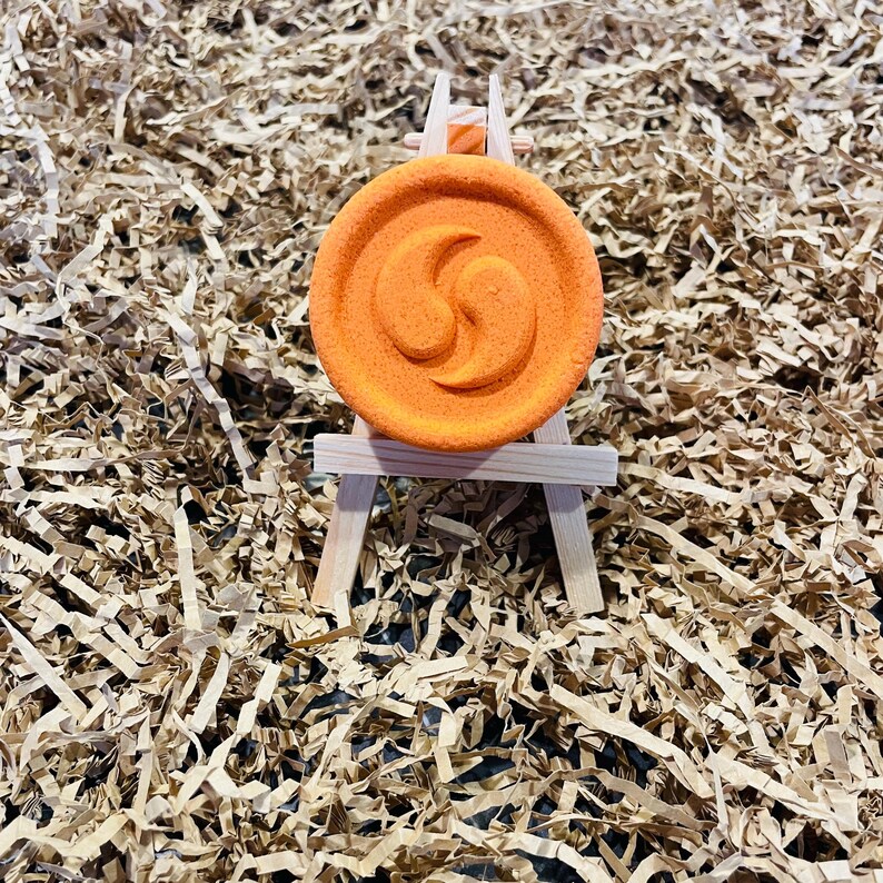 May include: An orange, circular object with a spiral design, displayed on a miniature wooden easel. The object is set against a backdrop of shredded brown paper, creating a textured and visually appealing presentation.