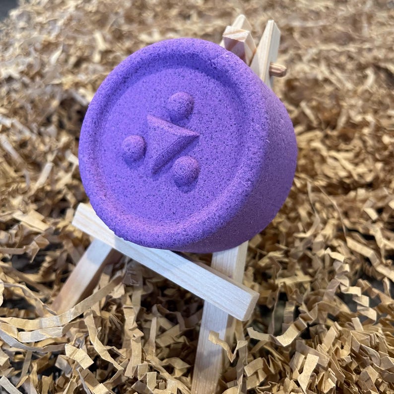 May include: A round, purple bath bomb with a geometric design of a triangle and two circles. The bath bomb is displayed on a small wooden easel, set against a background of brown paper shreds. The bath bomb is a vibrant shade of purple.