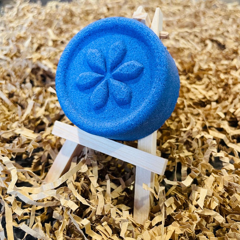 May include: A vibrant blue bath bomb with a floral design, resting on a miniature wooden easel. The bath bomb is round and features a raised flower pattern. The background is filled with shredded brown paper.