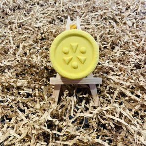 May include: A round, yellow bath bomb with a decorative design of triangles and circles, displayed on a miniature wooden easel. The bath bomb is set against a background of shredded brown paper.