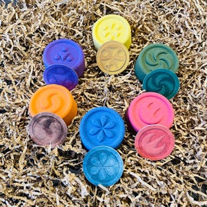 May include: A collection of colorful, round bath bombs in shades of yellow, purple, orange, blue, pink, and green. Each bath bomb has a matching, smaller, flat disc with a unique embossed design. The items are arranged on a bed of brown paper.