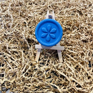 May include: A round, blue object with a flower design is displayed on a miniature wooden easel. The object is centered against a background of shredded brown paper. The flower design is embossed on the blue surface.