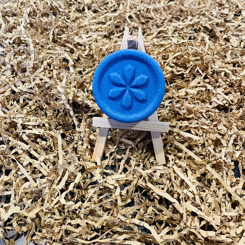 May include: A round, blue object with a flower design is displayed on a miniature wooden easel. The object is centered against a background of shredded brown paper. The flower design is embossed on the blue surface.