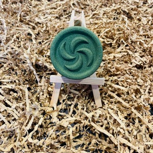 May include: A round, dark green object with a spiral design, displayed on a miniature wooden easel. The object is set against a backdrop of shredded brown paper, creating a textured and earthy aesthetic.
