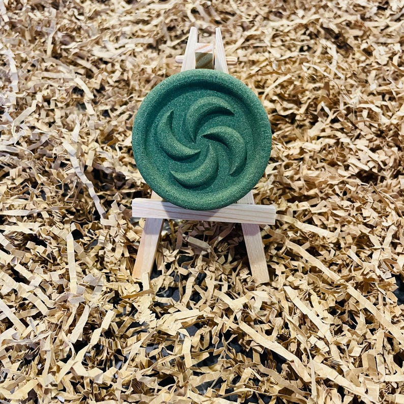 May include: A round, dark green object with a spiral design, displayed on a miniature wooden easel. The object is set against a backdrop of shredded brown paper, creating a textured and earthy aesthetic.