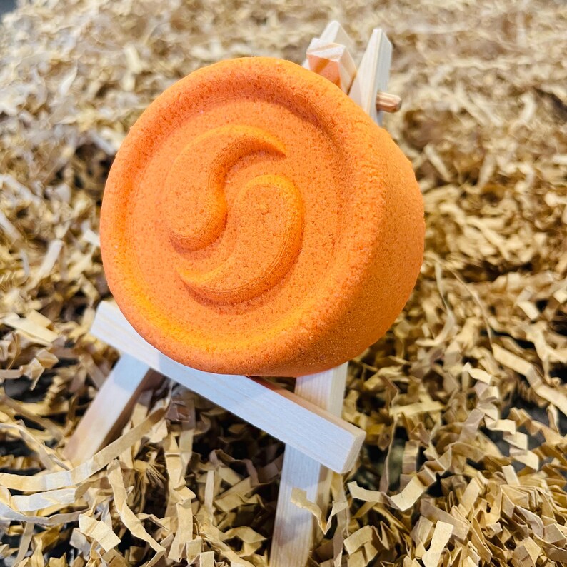 May include: An orange bath bomb with a spiral design, resting on a miniature wooden easel. The bath bomb is a vibrant shade of orange, and the easel is made of light-colored wood. The background is filled with brown paper shreds.