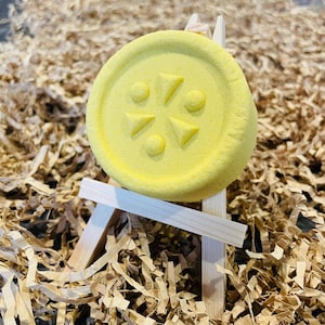 May include: A round, yellow bath bomb with a geometric design of triangles and circles. The bath bomb is displayed on a small wooden easel, set against a backdrop of shredded brown paper.