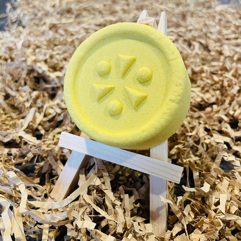 May include: A round, yellow bath bomb with a geometric design of triangles and circles. The bath bomb is displayed on a small wooden easel, set against a backdrop of shredded brown paper.