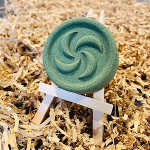 May include: A round, green bath bomb with a spiral design, displayed on a miniature wooden easel. The bath bomb is a muted green color and sits on a bed of brown paper shreds.