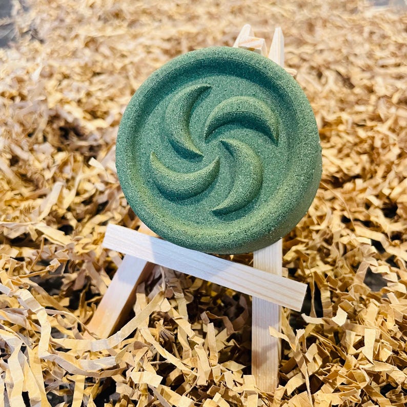 May include: A round, green bath bomb with a spiral design, displayed on a miniature wooden easel. The bath bomb is a muted green color and sits on a bed of brown paper shreds.