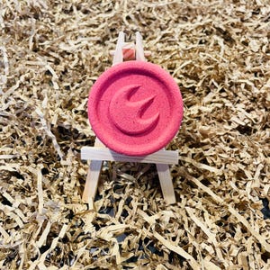 May include: A round, pink bath bomb with a carved design, displayed on a miniature wooden easel. The bath bomb has a textured surface and a stylized emblem. The background is filled with brown paper shreds.